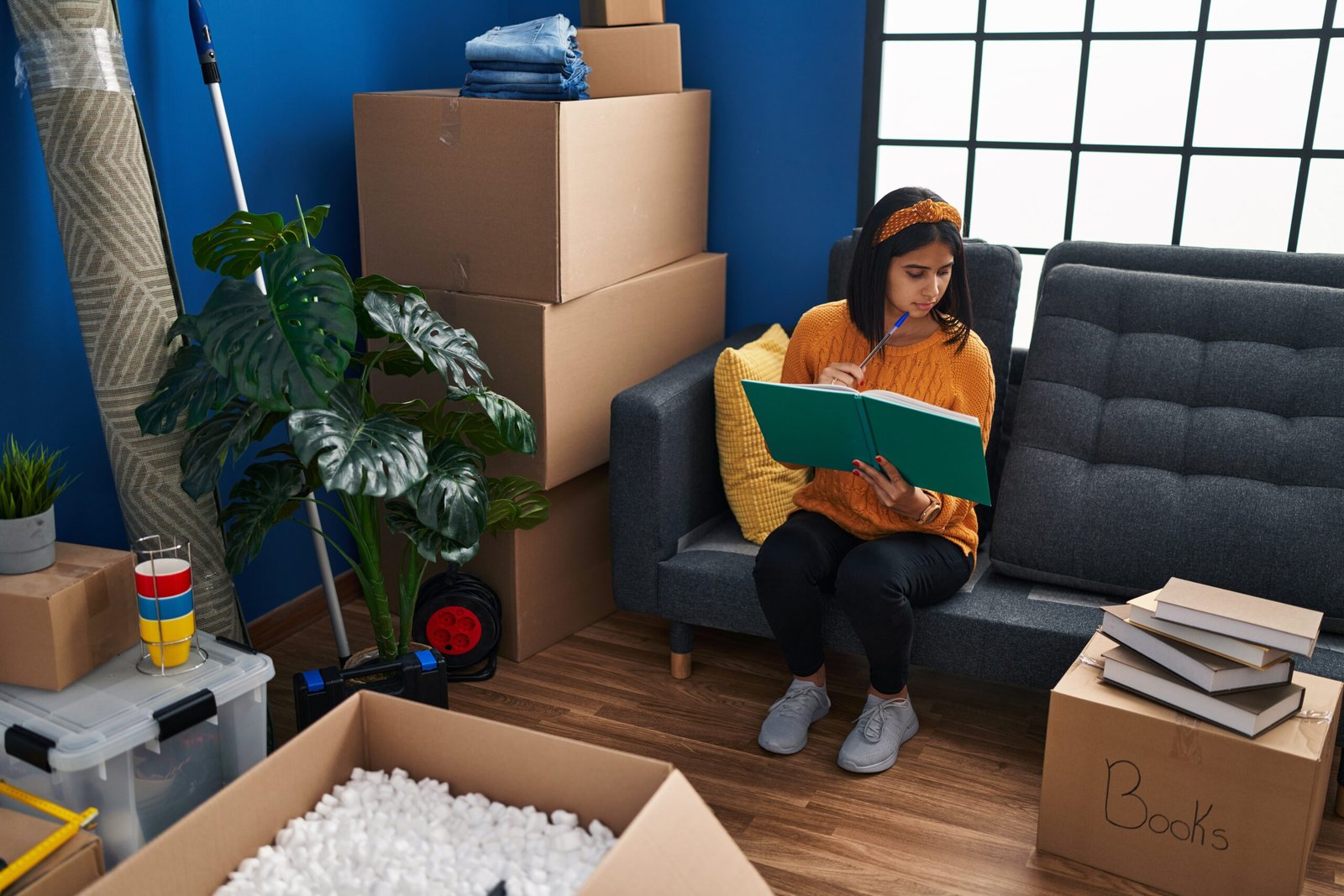 Young latin woman reading book sitting on sofa at new home