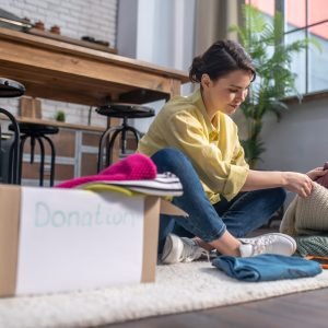 Smiling focused young dark-haired Caucasian woman seated on the floor selecting clothes for charitable causes